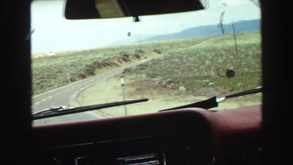 In-car 8mm view from a 1968 road trip, looking over a red dashboard toward a curving road in dry terrain.