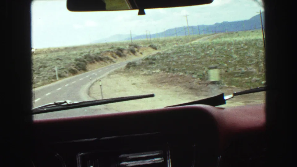1968 in-car travel footage looking over the dashboard at a quiet road bending through open, dry landscape.