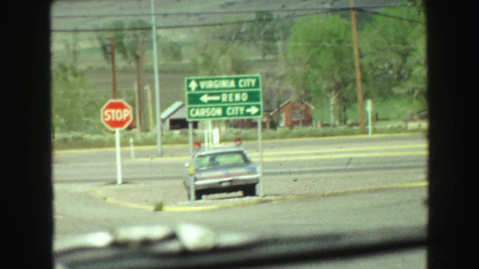 Road junction with a stop sign, an older car, and directional signs pointing toward Virginia City, Reno, and Carson City.