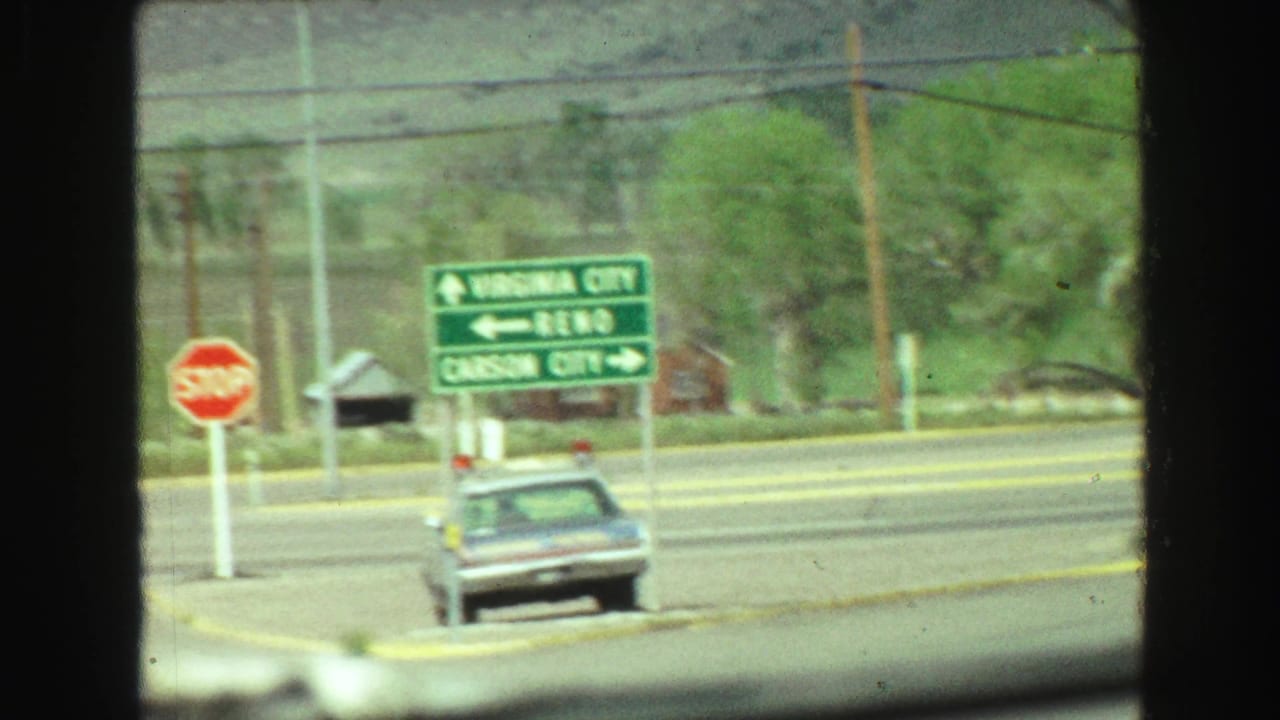 Roadside directional sign for Virginia City, Reno, and Carson City beside a stop sign and vintage sedan.