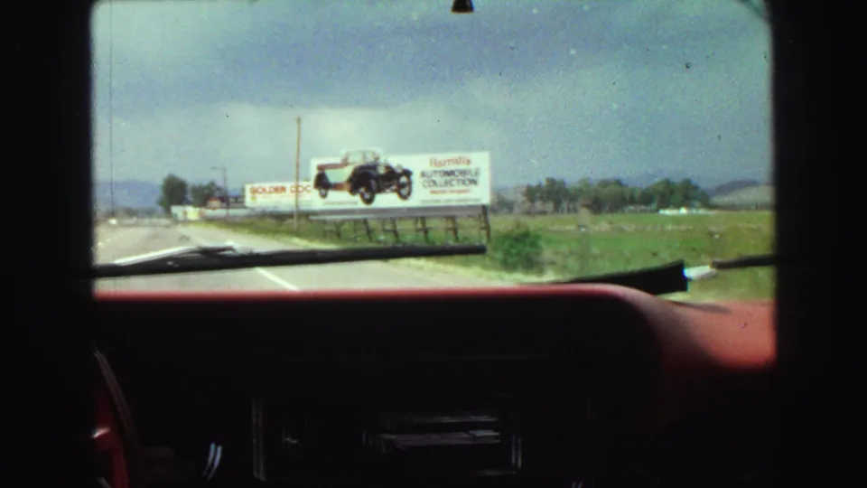From inside the car, the camera looks toward a roadside billboard for an automobile collection beyond the red dashboard and windshield.