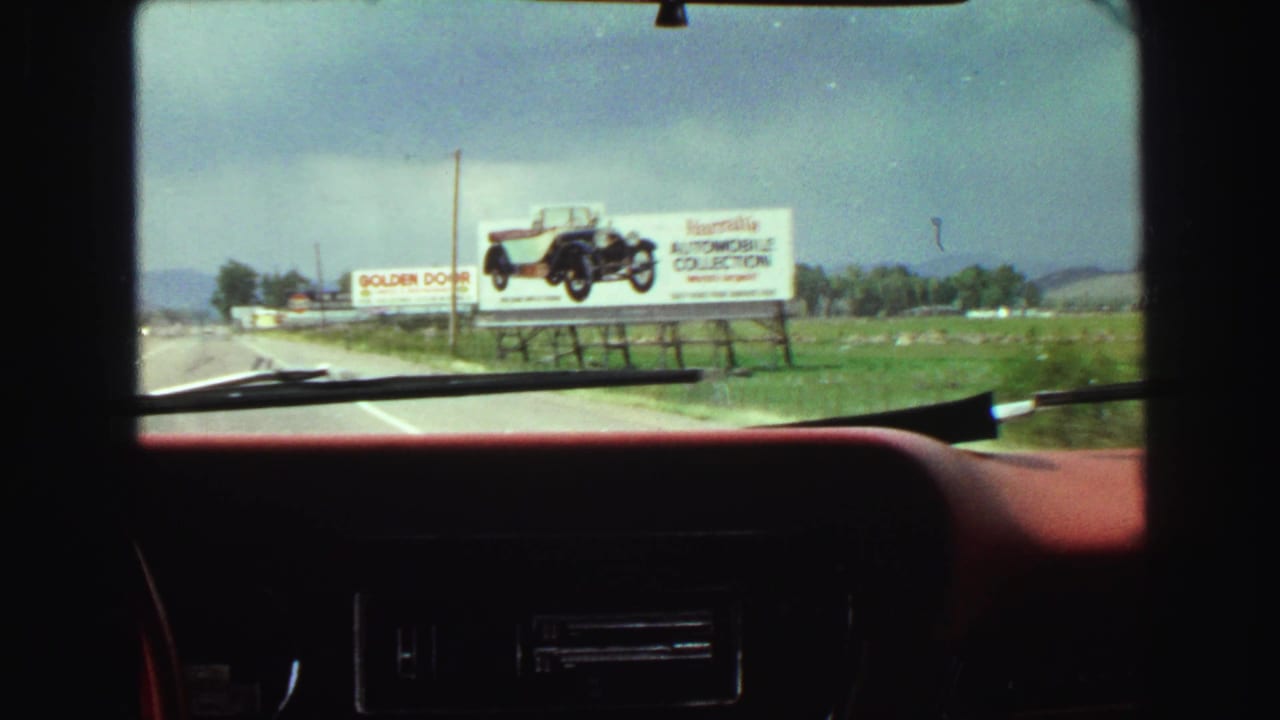 View through a car windshield toward a roadside billboard beside grass and distant hills.