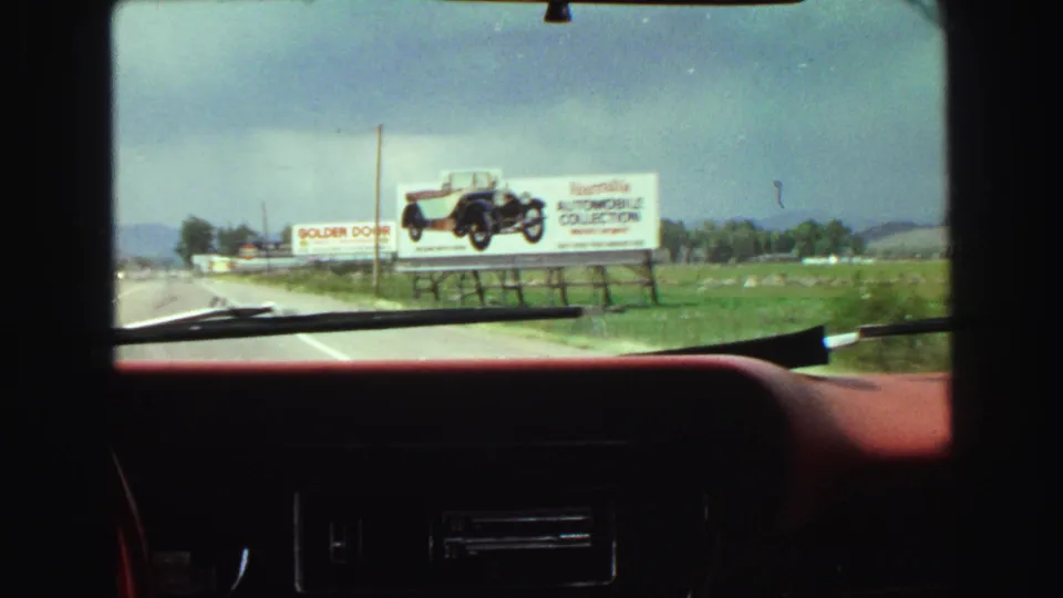 1968 8mm view from inside a red car near Vancouver, showing a roadside automobile billboard, grass, cloudy sky, and hills.