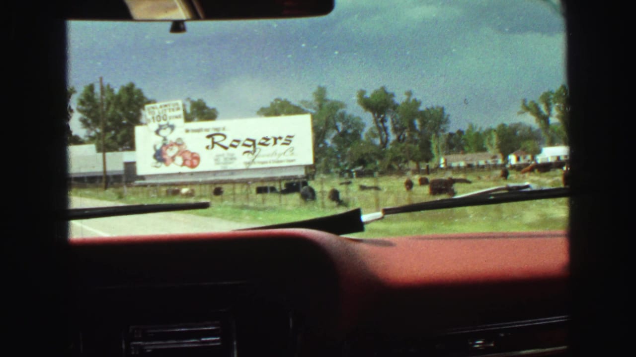 1968 Vancouver-area road footage from inside a red car, with a Rogers Jewelry billboard, open grass, trees, and dark animals visible.