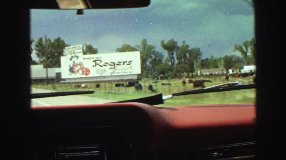 1968 Vancouver-area road footage from inside a red car, with a Rogers Jewelry billboard, open grass, trees, and dark animals visible.