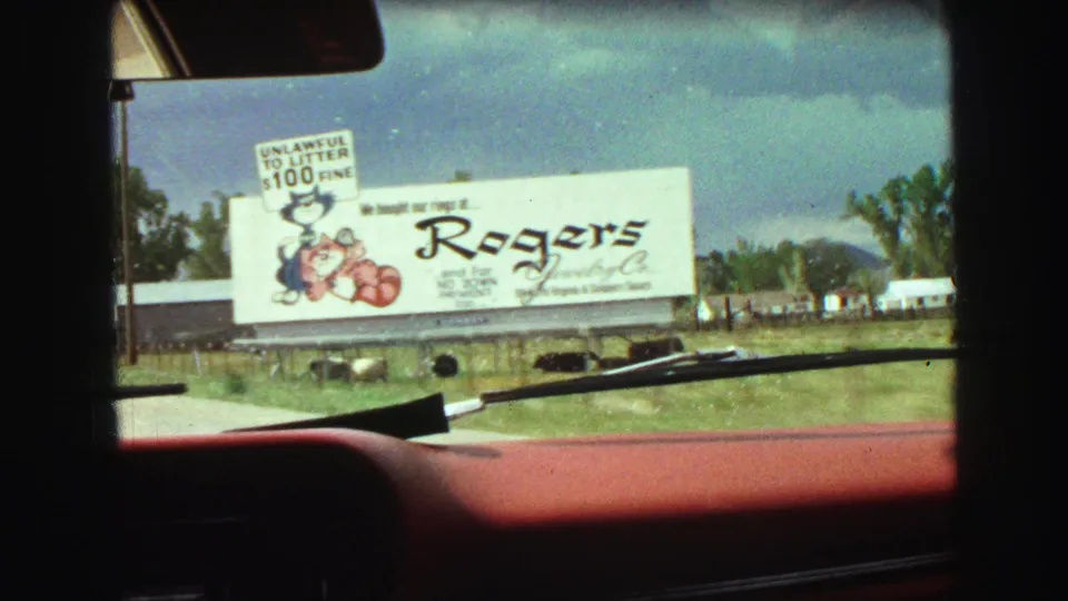 Closer 1968 8mm car-window view of a Rogers Jewelry billboard near Vancouver, with cartoon artwork, grass, trees, and windshield wipers.