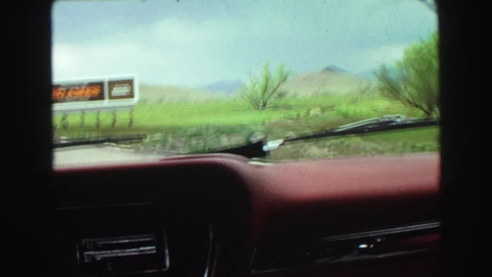 1968 Vancouver-area windshield view with red dashboard, roadside billboard, green grass, shrubs, cloudy sky, and distant hills.