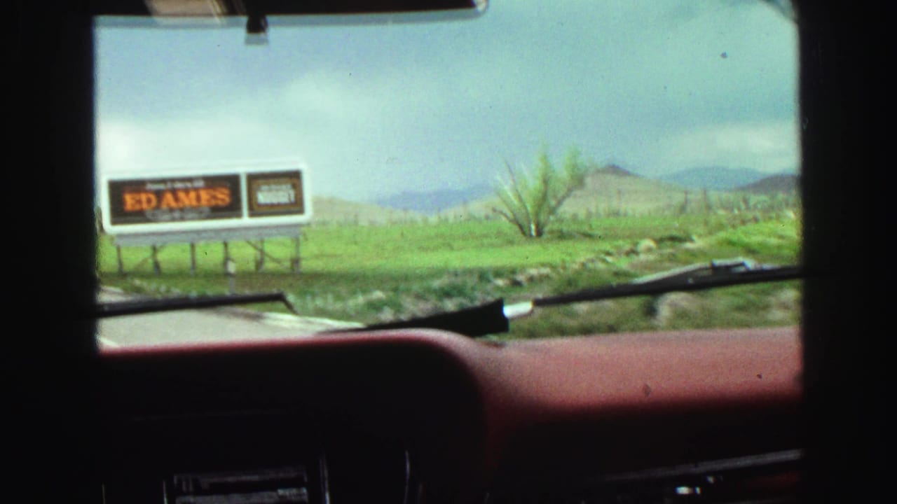 View through a car windshield toward an Ed Ames billboard beside green hills.