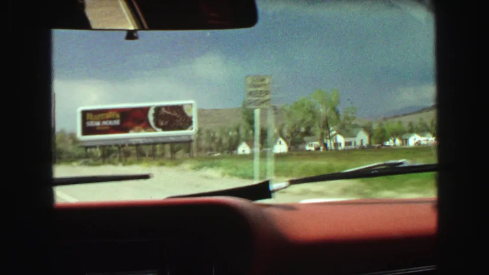1968 home-movie windshield view near Vancouver passing a Harrah's Steak House billboard, with trees and small houses in the background.
