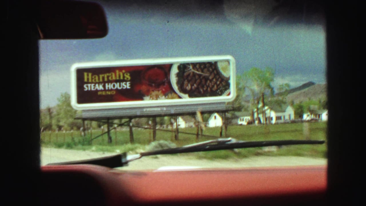 Windshield view of a roadside Harrah's Steak House billboard beside a road and open field.