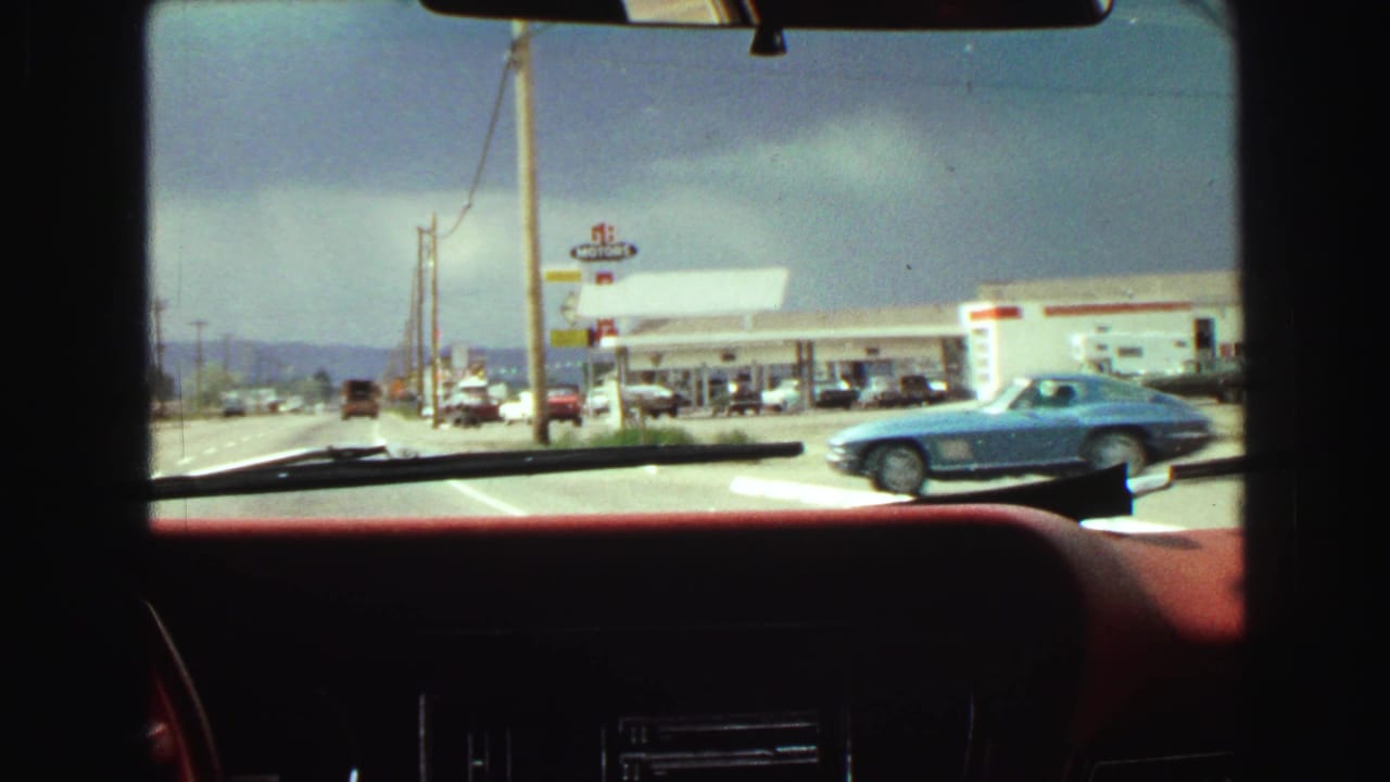 Roadside gas station area seen through a windshield, with a blue car passing at right.