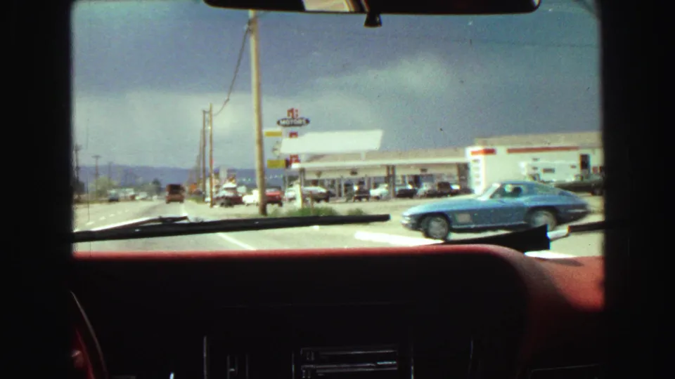 1968 home-movie view through a car windshield toward a gas-station frontage, road traffic, utility poles, and a blue sports car.