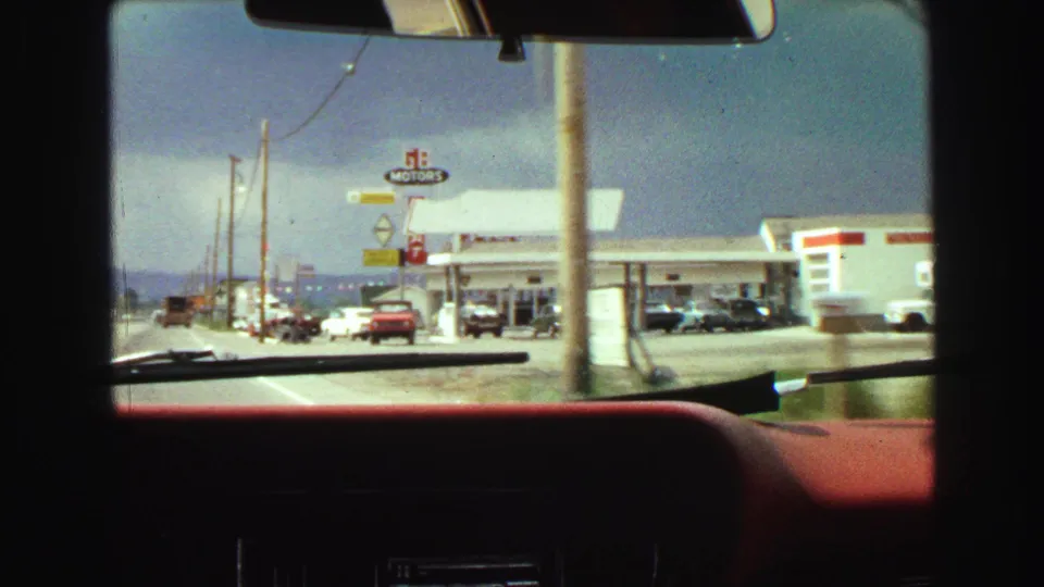 1968 windshield view of a roadside gas station, signs, parked vehicles, utility poles, and the car interior foreground.