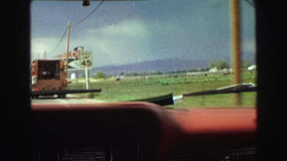 1968 in-car 8mm frame showing a roadside vehicle, a speed 45 sign, billboards, utility poles, fields, and mountains through the windshield.