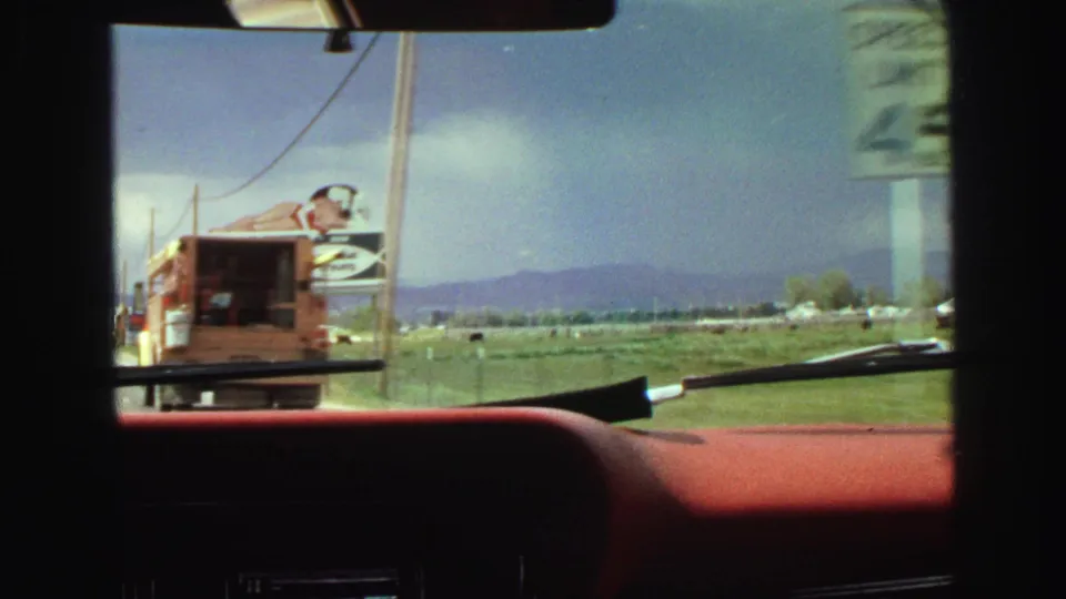 Windshield-framed 8mm view from a moving car in 1968 Vancouver, showing a roadside truck or trailer, billboard, fields, and distant mountains.