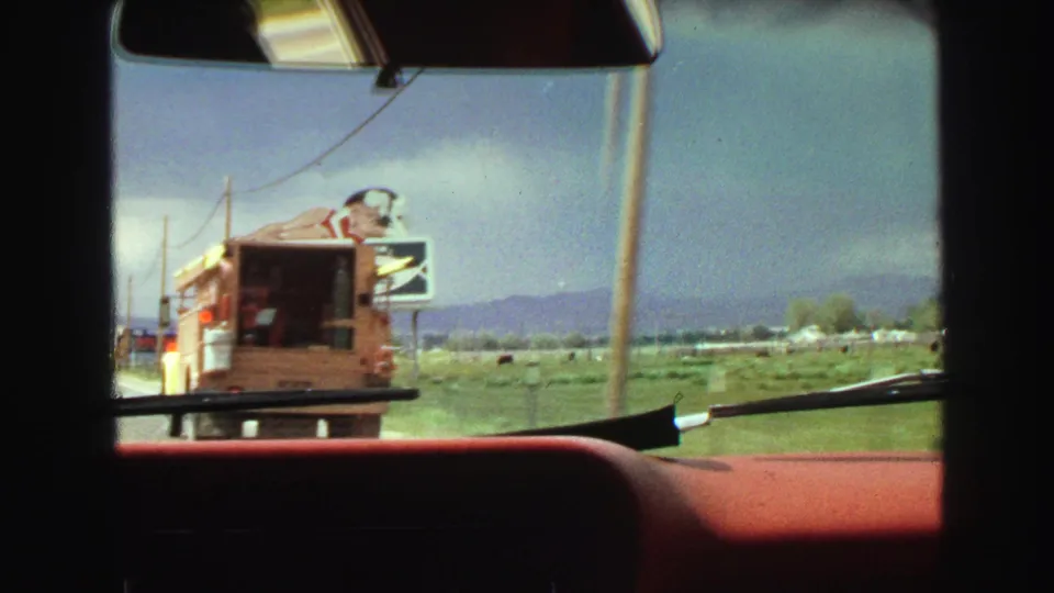 1968 home-movie view through a car windshield in Vancouver, with a red dashboard, wipers, a food truck, and distant roadside signs.