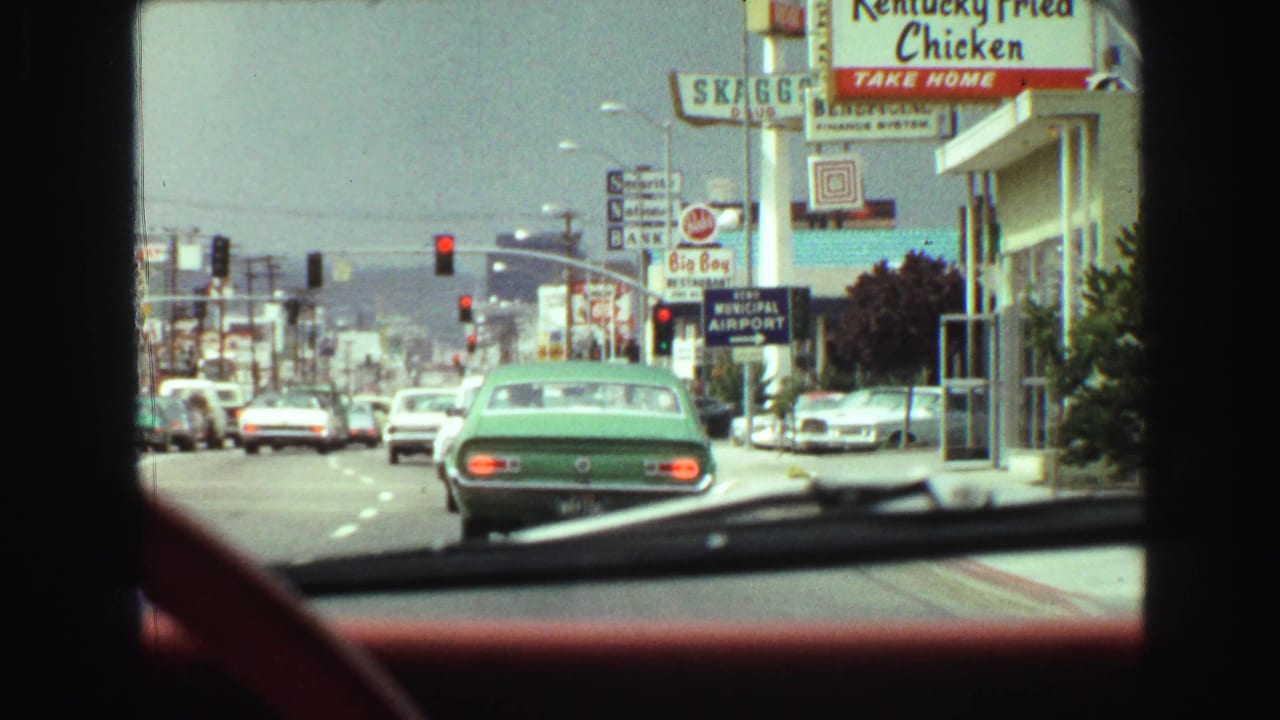 View through a car windshield of a busy street with a green car ahead and roadside signs.