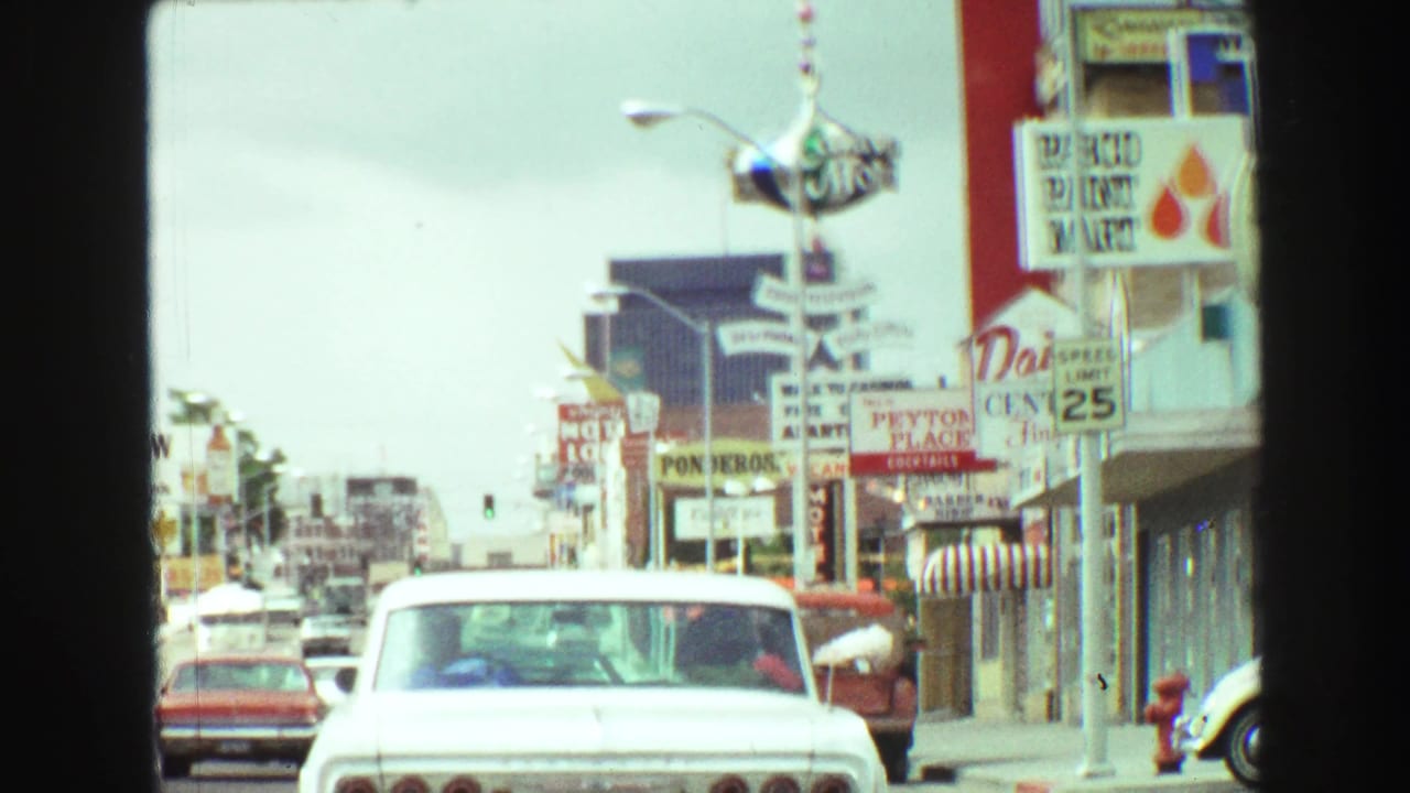 Street scene with a white classic car ahead and rows of motel, diner, and shop signs.