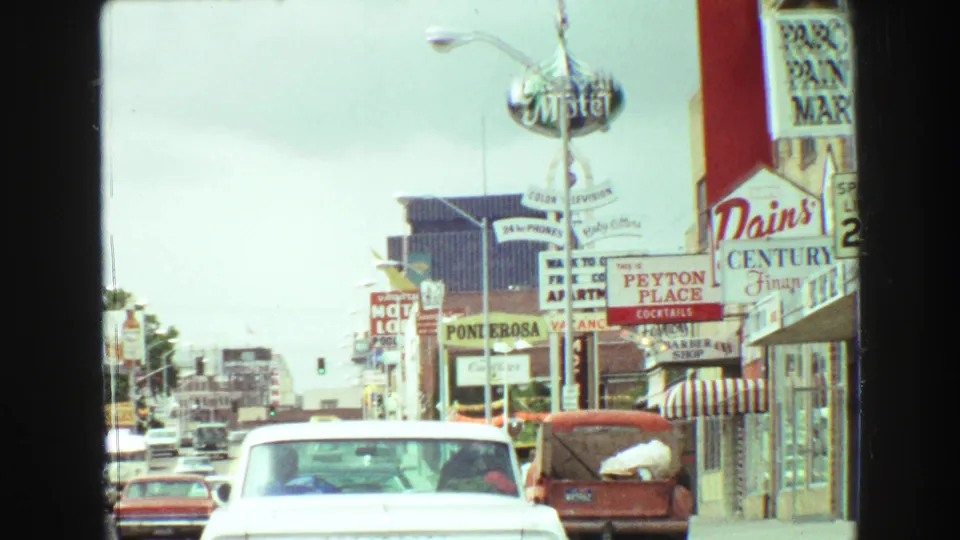 1968 Vancouver street footage showing a white classic car, dense roadside signs, storefront windows, and commercial buildings from an in-car viewpoint.