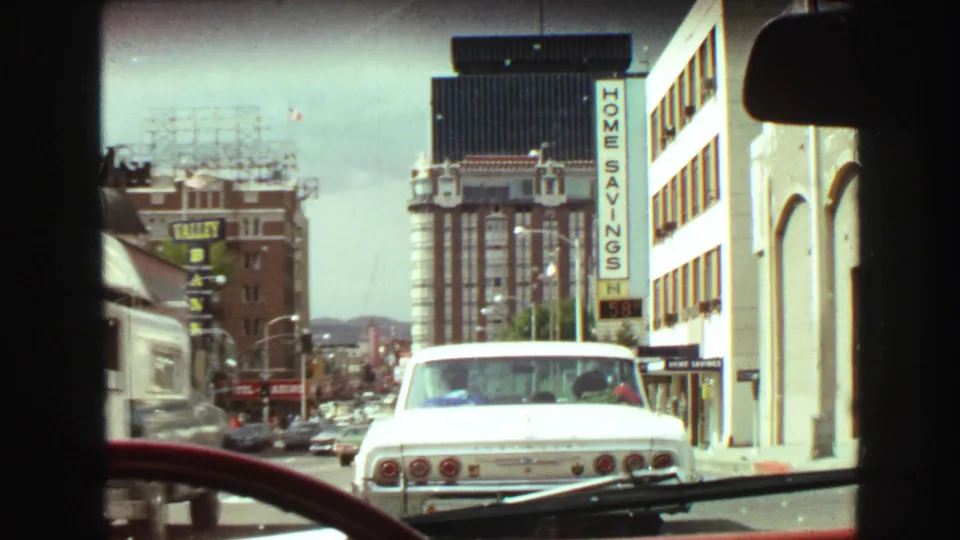 1968 Vancouver in-car street view with a white Chevrolet ahead, taller downtown buildings, a Home Savings sign, traffic, and distant hills.