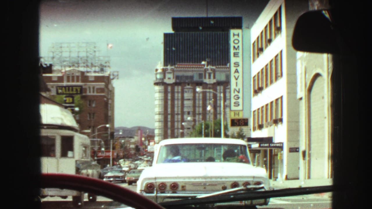 View through a vehicle windshield toward downtown traffic and a white car ahead.