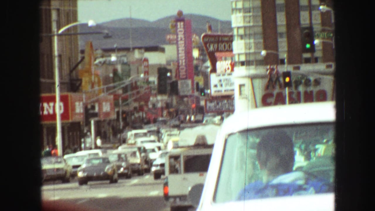 1968 Vancouver traffic scene with cars, storefront signage, traffic lights, distant hills, and a person visible in a foreground vehicle.