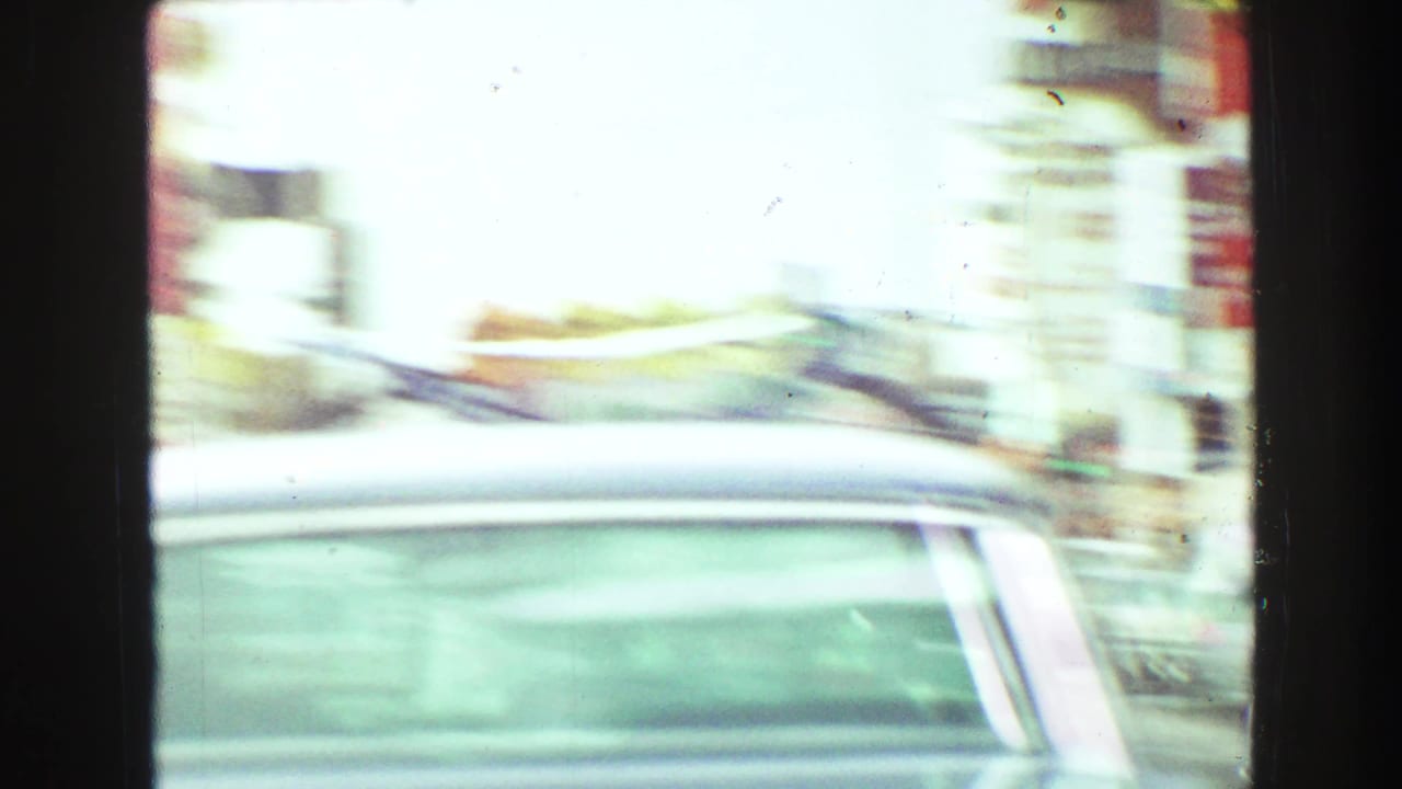 Motion-blurred 1968 home-movie frame of a passing car and commercial street, partially obscured by dark vehicle-window framing.
