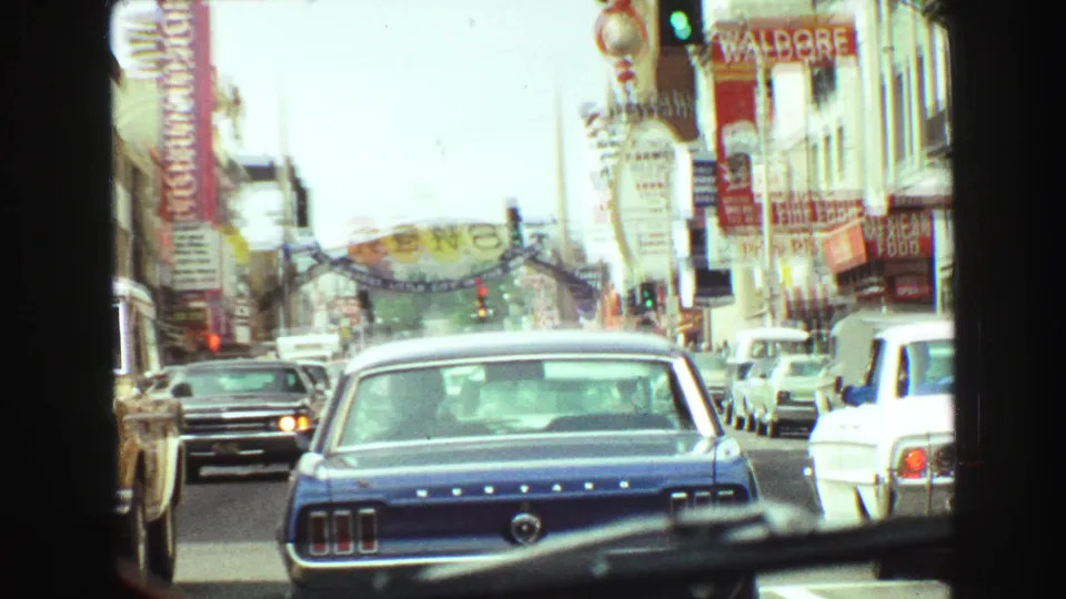 1968 8mm frame showing a blue Ford Mustang among traffic on a sign-lined commercial street, with a Reno arch visible ahead.