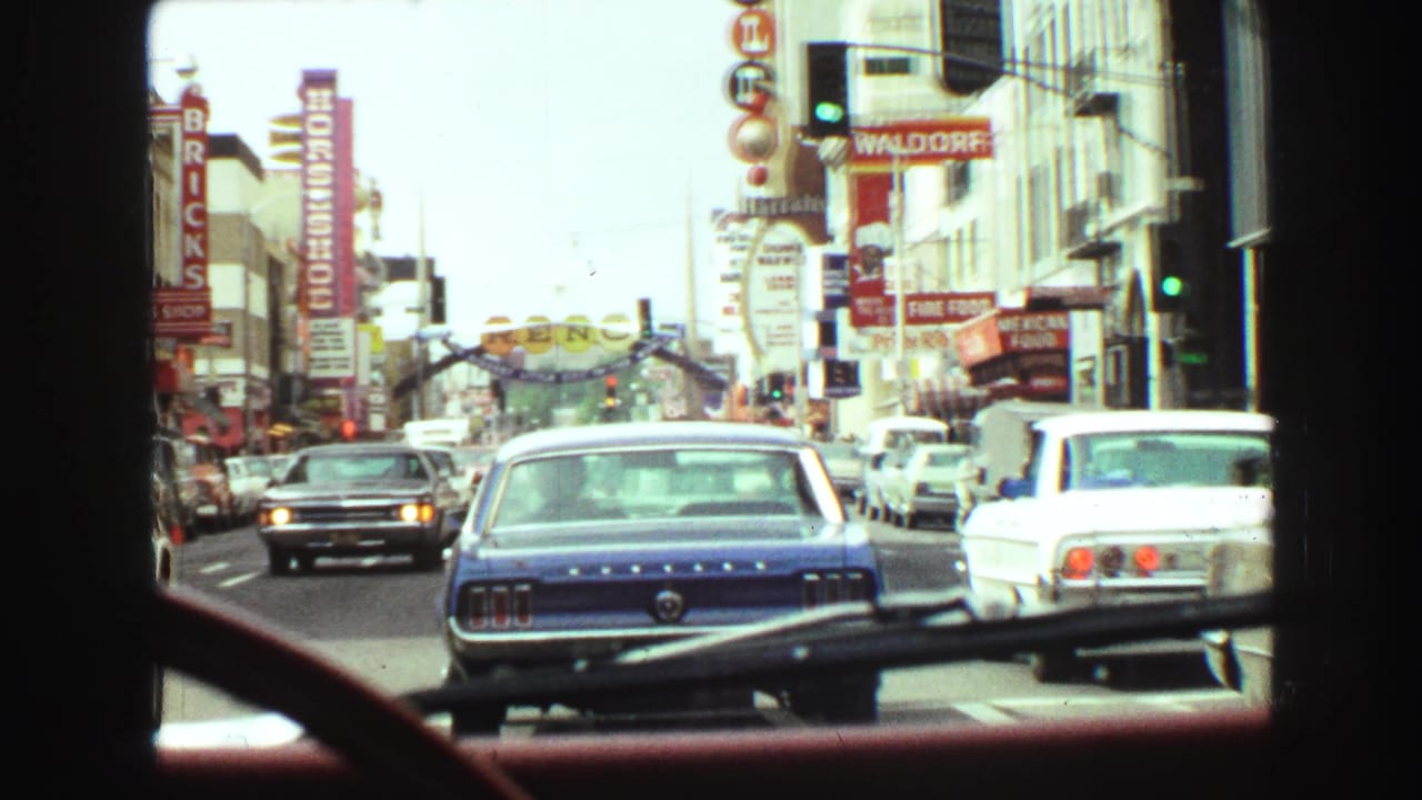 Traffic seen through a car windshield on a busy commercial street with vertical signs and classic cars.