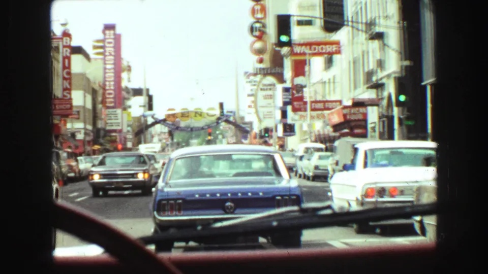 1968 Vancouver street-driving view through a windshield, with classic cars, traffic lights, and prominent hotel or theatre-style signs.