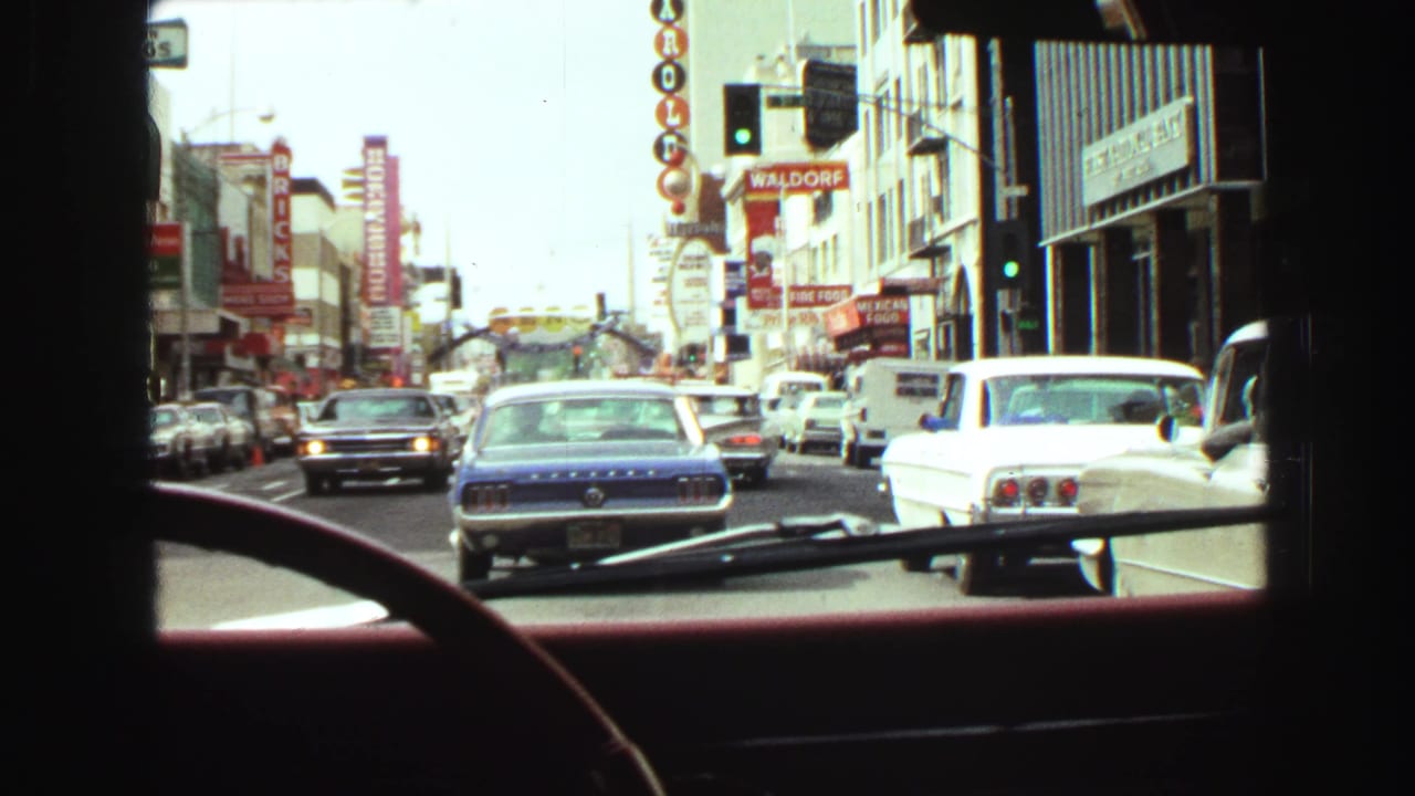 Forward street view from inside a car with a blue Mustang ahead and storefront signs along both sides.
