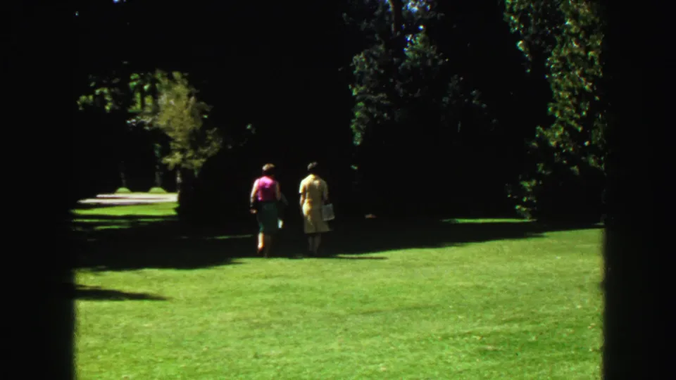 Park scene in Vancouver from the same 1968 home movie, with two distant people crossing a bright lawn near deep tree shade.