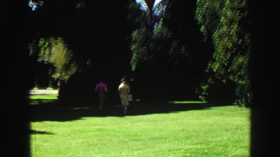 Late-1960s Vancouver park view with two distant pedestrians on a bright open lawn framed by tall, shadowed trees.