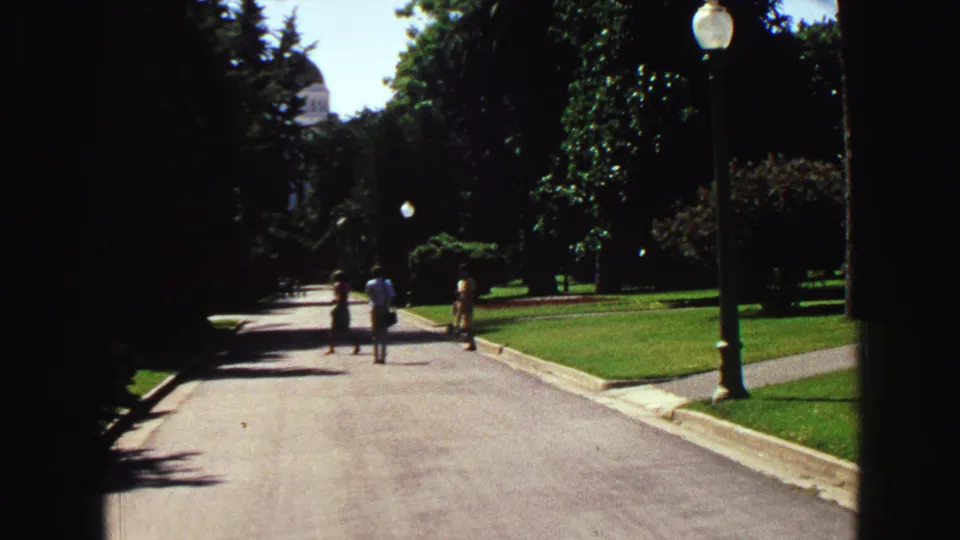 Distant visitors walk along a paved park road in 1968 Vancouver, bordered by lawns, trees, and lampposts.