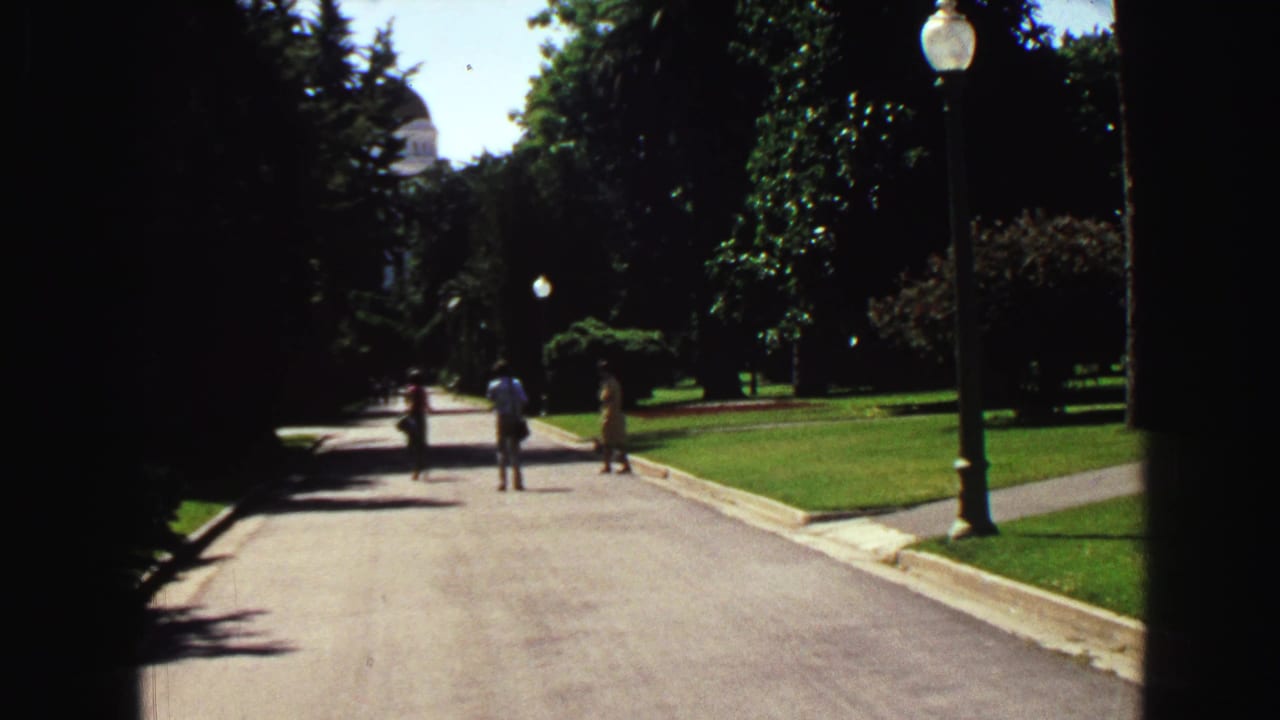 1968 Vancouver home-movie view down a sunlit park path with lawns, trees, globe lampposts, and distant walkers.