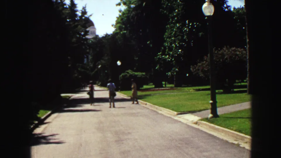 1968 Vancouver home-movie view down a sunlit park path with lawns, trees, globe lampposts, and distant walkers.