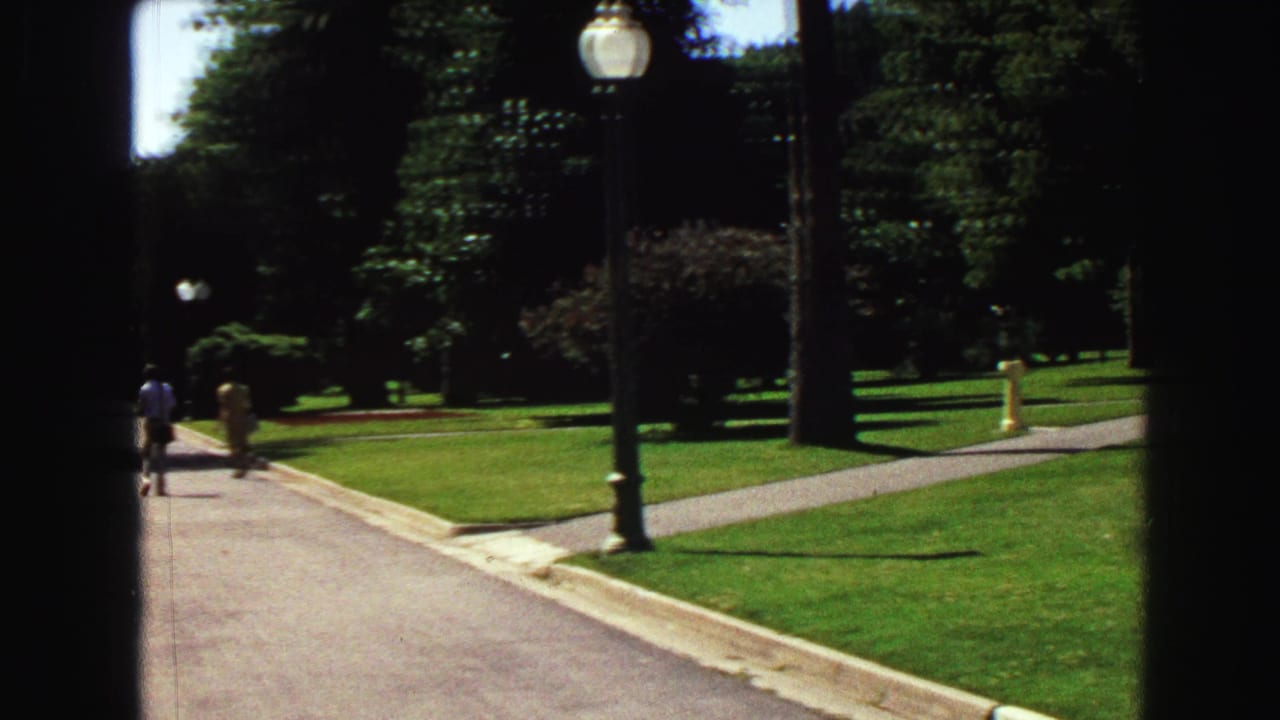 Park path curving past grass, trees, lampposts, and two distant walkers.