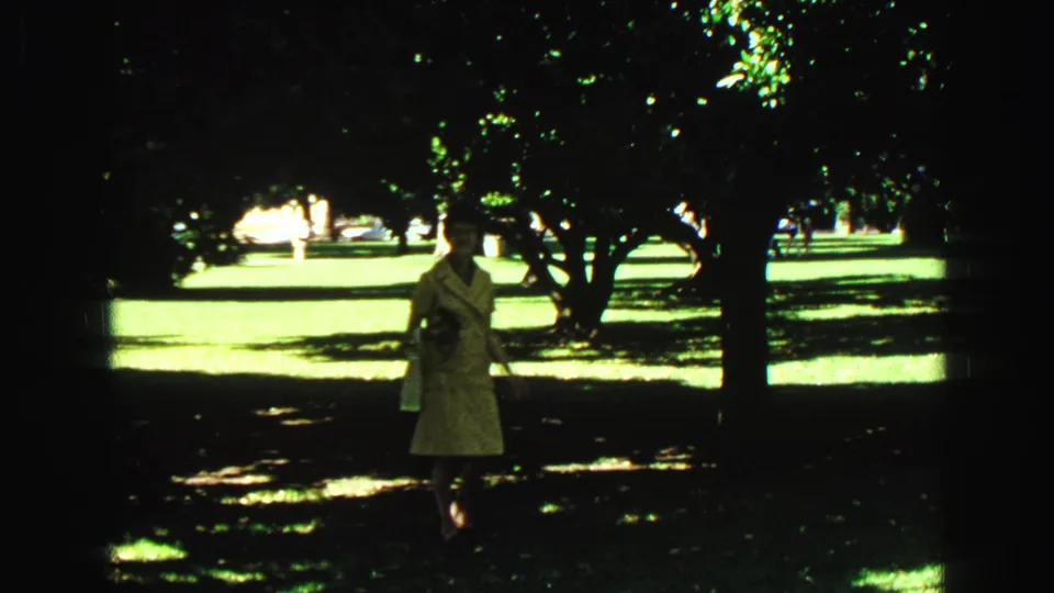 A woman in a light-colored dress walks through a shaded Vancouver park garden, with open grass and trees in the background.