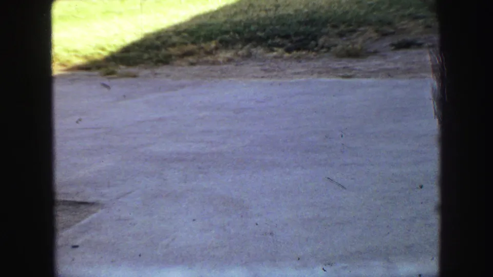 1968 Vancouver 8mm frame showing a paved surface, grassy rise, and shadowed border from a backyard or driveway area.