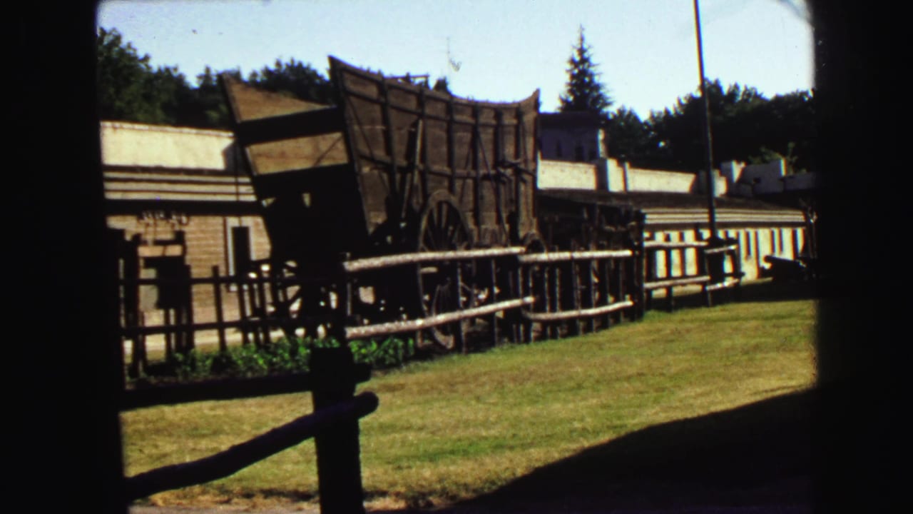Wooden wagon display beside low buildings and a lawn, seen through a dark opening.