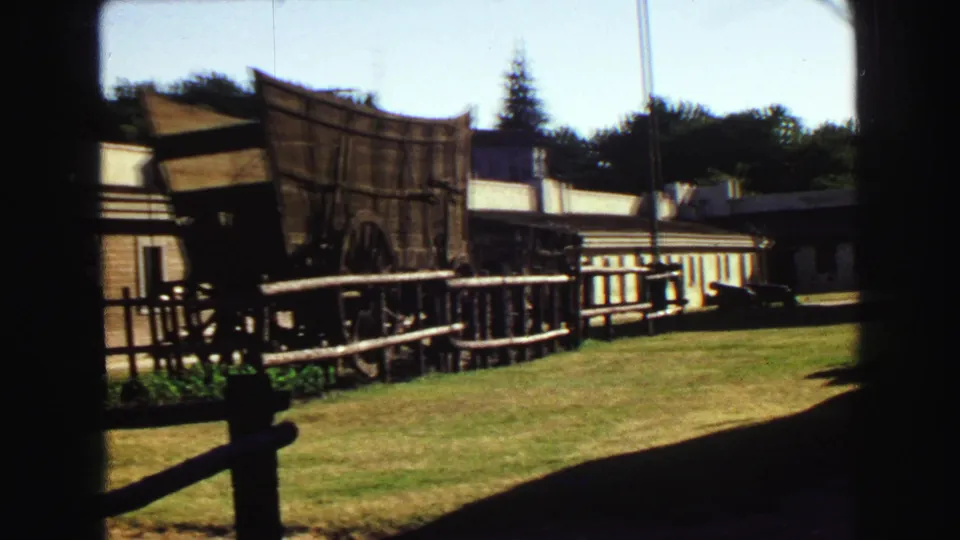 The camera frames the wooden wagon display, rail fence, grass, and long white building at a Vancouver heritage site in 1968 footage.