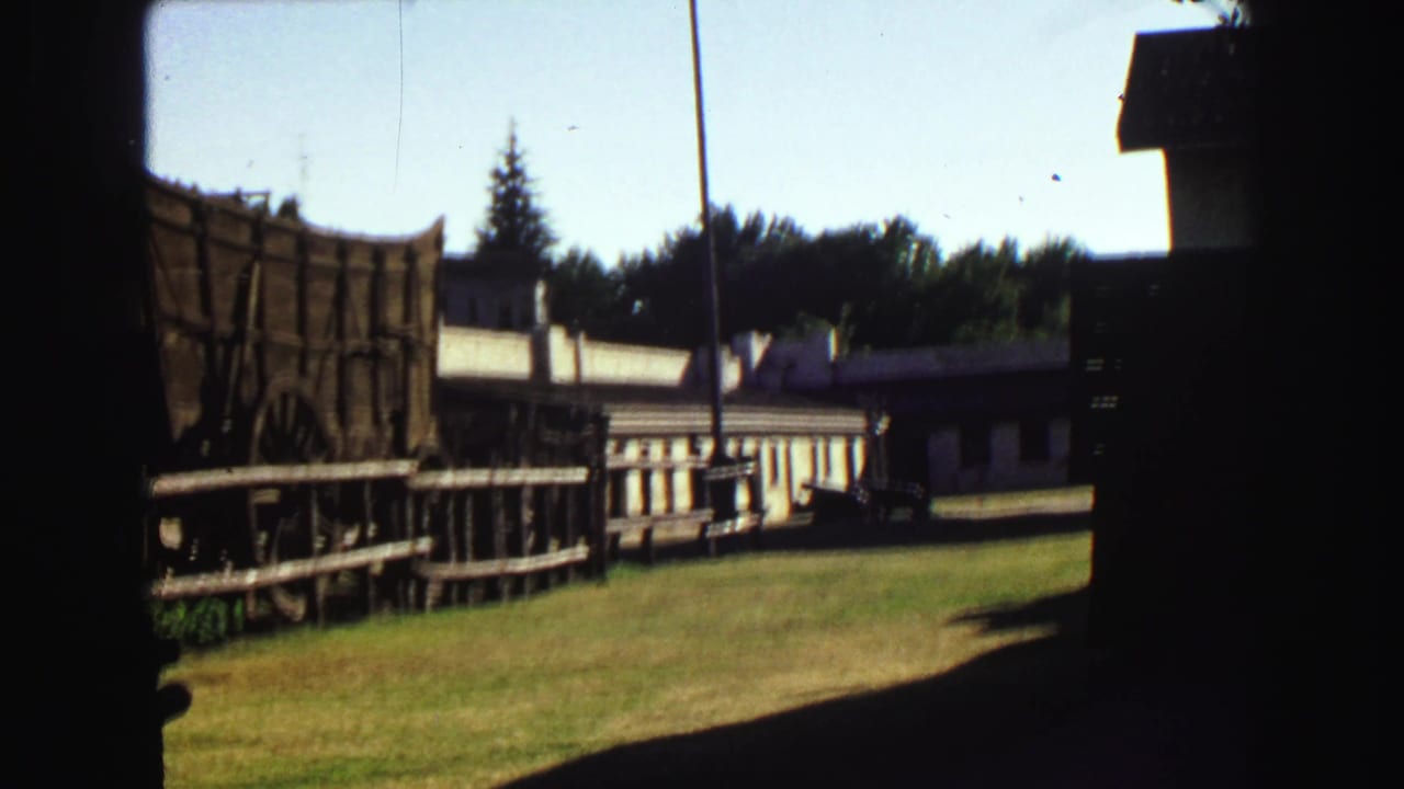 Wooden wagon and rail fence beside low historic buildings on a sunny lawn.