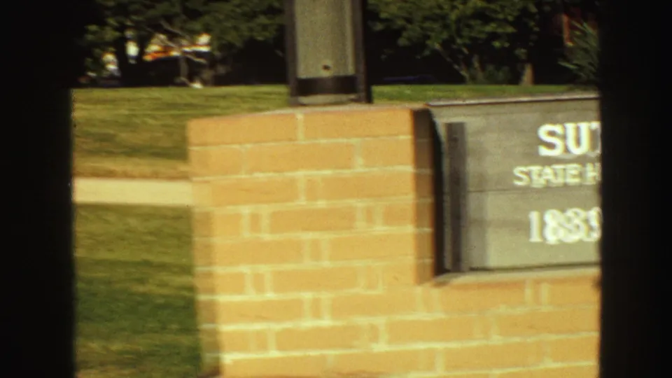 Partial 8mm frame of the Sutter's Fort monument sign and brick base, with lawn and plantings behind it.