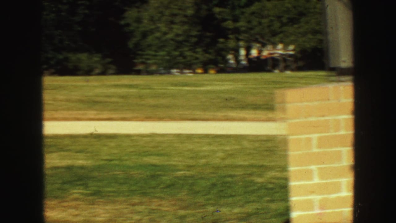1968 Vancouver home-movie frame showing a green lawn, pale walkway, brick column, and dark foreground framing from inside or near a doorway.