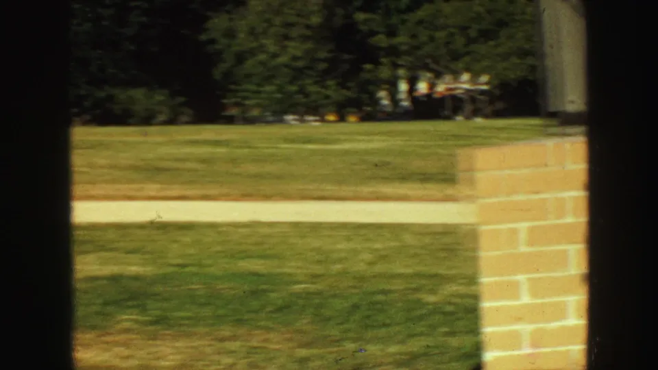 1968 Vancouver home-movie frame showing a green lawn, pale walkway, brick column, and dark foreground framing from inside or near a doorway.