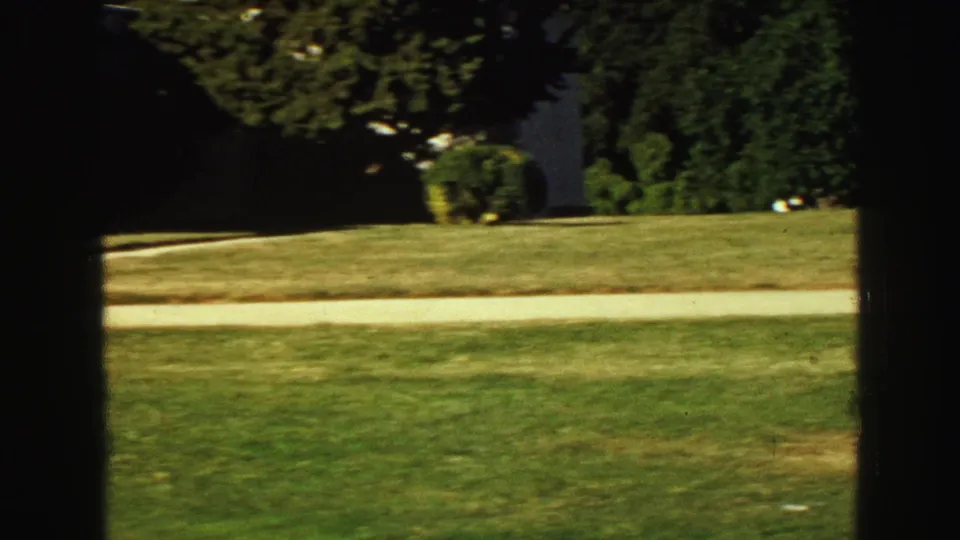 1968 Vancouver garden view with clipped grass, a light path, trees, shrubs, and a white wall partially blocked by dark foreground framing.