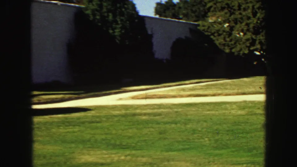 1968 Vancouver home-movie view of a white stucco wall beside a curving walkway, lawn, shrubs, and deep tree shadows.