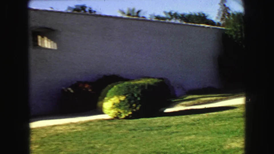 1968 Vancouver frame showing a white wall with a small arched window, rounded shrub, walkway, and sunlit lawn in a residential garden view.