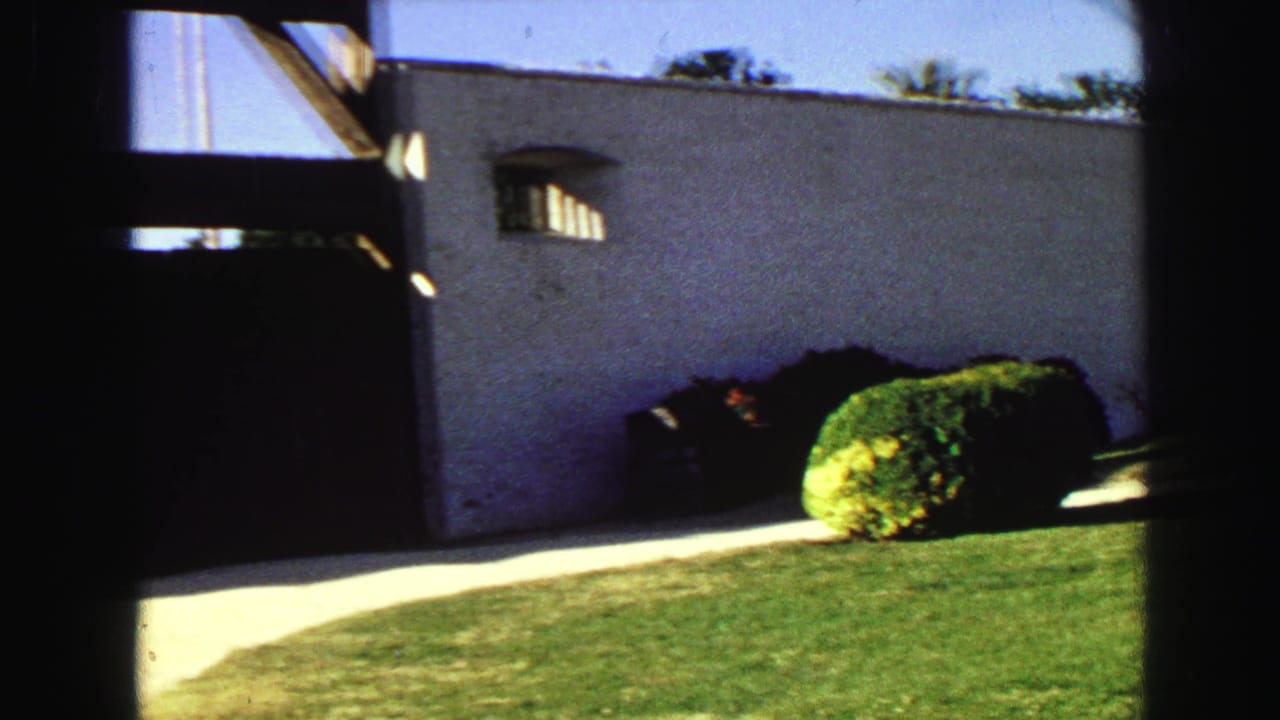 White stucco building with a dark wooden gate, small arched window, lawn, and shrubs in bright sun.