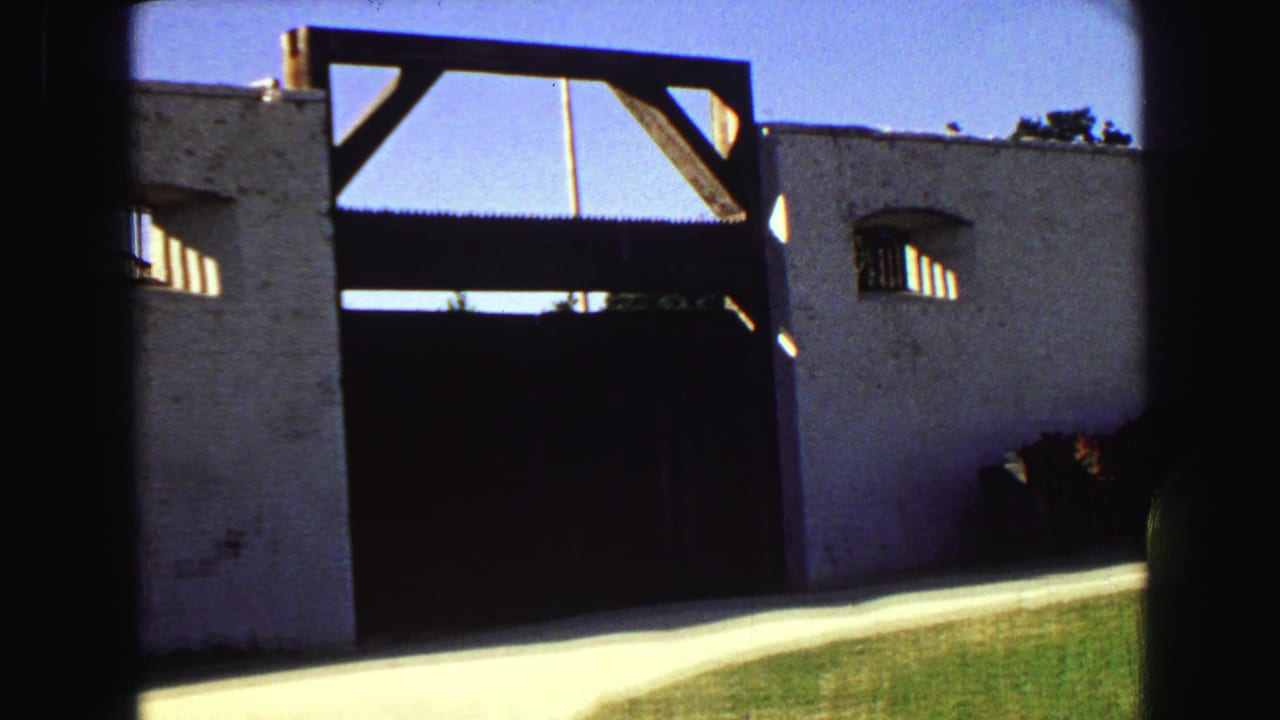 Closer view of a white stucco wall, large dark gate, wooden beam, small window, lawn, and sidewalk.